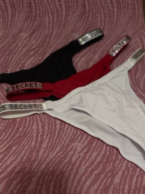 Victoria's Secret trosa 3-pack - Säljer ett 3-pack trosor från Victoria's Secret i färgerna svart, röd och vit. Alla har glittriga band med logotypen på sidorna. Materialet är mjukt och stretchigt, perfekt för en bekväm känsla och snygg look.