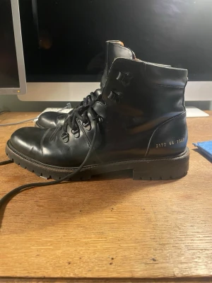 Svarta Common Prodjects läderkängor - Säljer ett par svarta Common Projects kängor i äkta läder med kraftig gummisula och snörning. Kängorna har metallhål för snören och detaljer i form av siffror på sidan. Perfekta för dig som gillar en robust och stilren look. Priset är inte hugget i sten