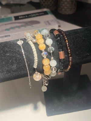 Flera armband med pärlor och detaljer - Säljer ett set med flera armband i olika stilar. Armbanden har pärlor i gult, vitt, svart och brunt samt inslag av silverfärgade detaljer och kristalliknande pärlor. Några armband har metallinslag och ett har en tunn kedja med hjärtdetalj. Perfekt för dig som gillar att mixa och matcha. kan diskutera pris