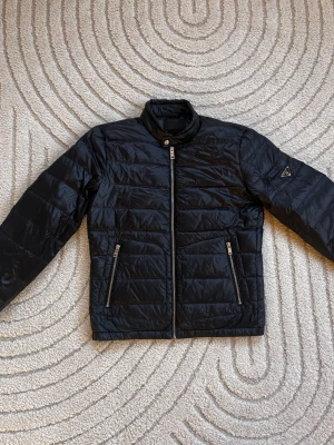 Svart dunjacka från Prada - Snygg svart dunjacka från Prada med quiltad design och raka linjer. Jackan har två dragkedjeförsedda fickor framtill och en hög krage. Klassisk Prada-logga på vänster ärm. Perfekt för kalla vinterdagar.