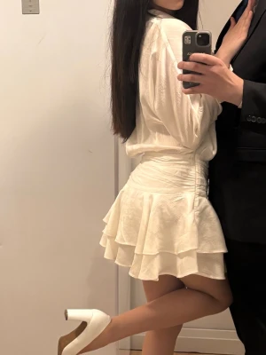 Vit omlottklänning med volang - Kort vit omlottklänning med volangdetaljer och markerad midja. Klänningen har långa ärmar och en flowy kjol med lager på lager-effekt. Perfekt för dig som gillar en feminin och trendig stil.