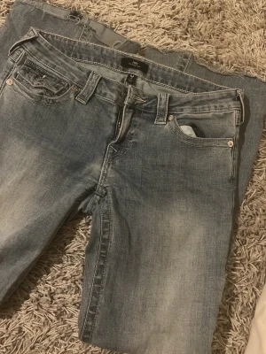 True Religion Joey Low Rise Flare Jeans - Snygga blå jeans från True Religion, modell Joey Low Rise Flare. Jeansen har låg midja, utsvängda ben och klassisk femficksdesign. Tillverkade i bomull med dragkedja och knapp framtill. Perfekta för en avslappnad och trendig look. Har lite missfärgning där nere (kolla sista bilden) därav priset är billigare 
