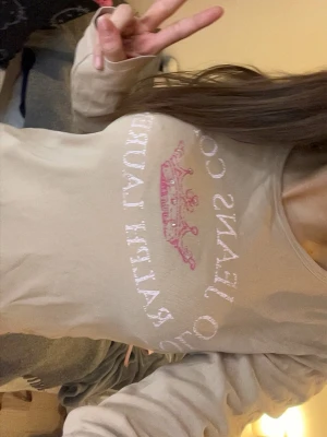 Y2K Beige långärmad topp Ralph Lauren - Snygg beige långärmad topp från Ralph Lauren Jeans Company med ett rosa kronmotiv och text framtill. Toppen har rund halsringning och är gjord i mjukt bomullsmaterial. Perfekt för en chill och trendig look.