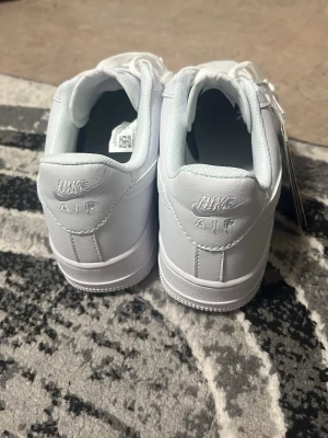 Nike Air Force 1 vita sneakers - Klassiska helvita Nike Air Force 1 sneakers i lågt utförande. Skorna har en stilren design med perforerad tå, vita skosnören och Nike-logga broderad på hälen. Tillverkade i slittåligt läder med en robust sula för extra komfort.