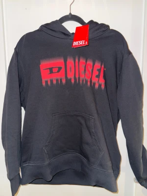 Svart hoodie från Diesel med röd logga - Svart hoodie från Diesel med stor röd logga och grafiskt tryck framtill. Klassisk känguruficka och huva. Mjuk bomullsblandning som är skön att ha på sig. Perfekt för en avslappnad streetstyle-look.