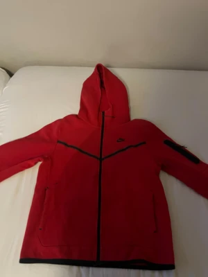 Röd Nike hoodie med dragkedja - Röd hoodie från Nike med hel dragkedja framtill, svarta detaljer och fickor på sidorna. Huva och en extra ficka med dragkedja på ena ärmen. Perfekt för en sportig och avslappnad stil.