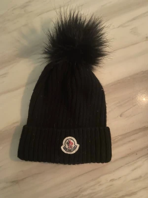 Svart Moncler mössa med pälsboll - Snygg svart ribbstickad mössa från Moncler med stor fluffig pälsboll på toppen. Mössan har uppvikt kant och klassisk Moncler-logga framtill. Perfekt accessoar för kalla dagar och en stilren look.