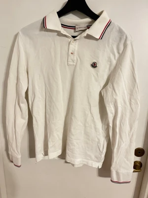 Moncler piké - Vit långärmad pikétröja från Moncler med klassisk krage och knäppning framtill. Tröjan har Moncler-logga på bröstet och röda samt marinblå detaljer på krage och ärmslut. Tillverkad i mjuk bomull för en clean och stilren look.