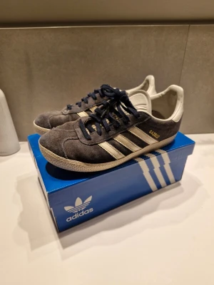 Adidas Gazelle mörkblå sneakers - Snygga Adidas Gazelle sneakers i mörkblå mocka med vita detaljer och klassiska tre ränder på sidan. Skon har guldfärgad Gazelle-text, vita sulor och mörkblå snören. Perfekt för dig som gillar retrostil och vill ha något tidlöst från Adidas.