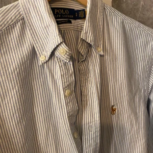 Randig skjorta från Ralph Lauren - Säljer en stilren blå- och vitrandig skjorta från Ralph Lauren. Skjortan har långa ärmar och klassiska knappar framtill. Den är broderad med den ikoniska loggan på bröstet. Perfekt för en snygg och avslappnad look. Storlek S och i extremt bra skick 