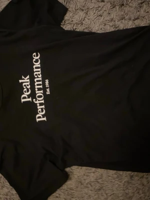 Svart Peak Performance t-shirt - Svart t-shirt från Peak Performance med vit logga och texten 'Est. 1986' på bröstet. Klassisk passform med rund hals och korta ärmar. Perfekt för en stilren och avslappnad look.