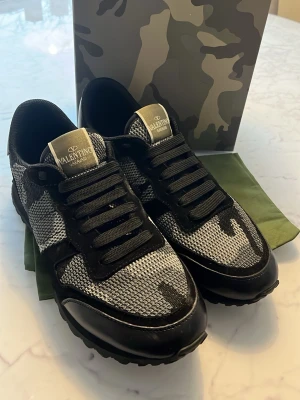Valentino camo sneakers svart/grå - Snygga sneakers från Valentino i svart och grått med camomönster. Ovandelen är i mesh och läder, med svarta skosnören och robust sula. Märkeslogga på plösen och cool streetstil. Perfekta för dig som vill sticka ut med exklusiva detaljer.
