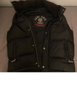 Svart dunväst från GANT - Snygg svart dunväst från GANT med hög krage och två stora fickor med dragkedja. Västen har quiltad design och tryckknappar samt dragkedja framtill. Perfekt för lager-på-lager och streetwear-stil.