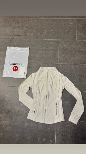  Vit Lululemon Define Jacket -                                                    Snygg och stilren träningsjacka från Lululemon i klassiskt svart med exklusiva guldfärgade dragkedjor som ger en lyxig touch ✨ Jackan är figurnära med smarta sömmar som framhäver silhuetten på ett väldigt smickrande sätt.  Perfekt för träning, yoga, gym eller som en sportig vardagsjacka. Materialet är stretchigt, följsamt och bekvämt – precis den höga kvalitet som Lululemon är kända för. Två praktiska fickor med drag