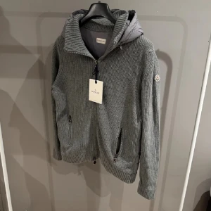 Grå Moncler cardigan -   Säljer en riktigt snygg och bekväm jacka från Moncler. Grå stickad modell med vadderad huva, perfekt för vår/höst eller mild vinter. Väldigt skön på och lätt att matcha till det mesta. Kvitto medföljer och alla tags!  • Dragkedja framtill • Fickor med dragkedja • Moncler-logga på ärmen • Original – tag kvar