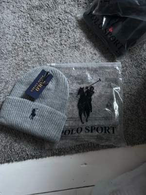 Grå ribbstickad mössa Polo Ralph lauren  - Snygg grå ribbstickad mössa från Polo Ralph Lauren  med klassisk broderad logga i mörkblått framtill. Mössan är i mjukt material och har en bred uppvikt kant för extra stilpoäng. Perfekt accessoar för kalla dagar.