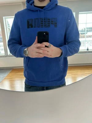 Blå hoodie från Puma - Snygg blå hoodie från Puma med svart logga på bröstet. Klassisk modell med huva och känguruficka framtill. Perfekt för en avslappnad och sportig stil. Mjuk och skön bomullsblandning.