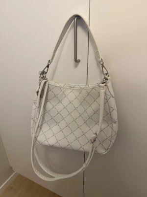 Vit axelväska från Tamaris  - Snygg vit axelväska från Tamaris i konstläder med rutmönster i grått. Väskan har både kort och lång axelrem samt silverfärgade metalldetaljer och en rund logga i metall. 