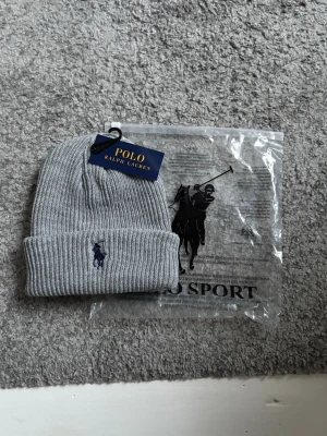 Grå ribbstickad mössa Polo Ralph Lauren - Snygg grå ribbstickad mössa från Polo Ralph Lauren med klassisk uppvikt kant och broderad marinblå logga framtill. Tillverkad i mjukt material som håller dig varm och passar perfekt till höstens och vinterns outfits.
