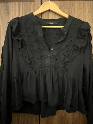 Svart blus med broderade detaljer - Svart blus från Bik Bok med långa ärmar, volanger och broderade blommönster framtill och på ärmarna. Blusen har knappar vid halsen och en lös, flowy passform. Perfekt för dig som gillar romantiska detaljer och en avslappnad stil.