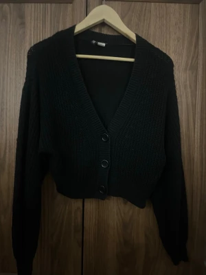 Svart stickad kofta - Snygg svart stickad kofta med v-ringning och tre stora knappar framtill. Koftan har lång ärm och ribbade muddar vid ärmslut och nederkant. Perfekt att slänga över en t-shirt eller linne för en chill look.