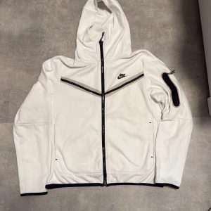 Vit Nike hoodiejacka med svarta detaljer - Vit hoodiejacka från Nike med svarta detaljer och dragkedja hela vägen. Jackan har en svart Nike-logga på bröstet, svarta linjer över bröstet och en ficka med dragkedja på ena ärmen. Tillverkad i 100% bomull och har huva.det svarta hade gåt av och jag fixade dden den är inte synlig om du inte zoomar in