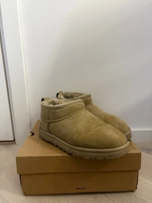 Beige UGG i mocka - Mysiga beige stövlar från UGG i mjuk mocka med fårskinnsfoder. Klassisk låg modell med rund tå och grov sula för extra komfort. Perfekta för att hålla fötterna varma hemma eller ute. UGG-logga bak på hälen och praktisk hälflik.