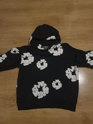 Denim Tears Mörkblå - Säljer en mörkblå hoodie med stora vita blommönster över hela plagget. Hoodien har en klassisk huva och ribbade muddar vid ärmslut och nederkant. Perfekt för dig som vill sticka ut med en unik och trendig stil.