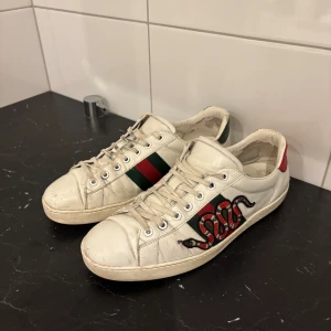 Orm broderad sko - Sneakers i vitt skinn med klassiska gröna och röda ränder på sidan, broderad orm och metallic-detaljer i rött och grönt på hälen. Skorna har snörning, rund tå och platt sula. Perfekt för dig som vill sticka ut med lyxig streetstyle. gucci ace