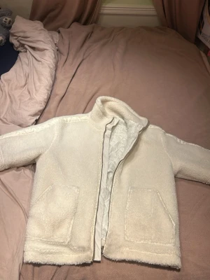 Vit teddyjacka med dragkedja - Mysig vit teddyjacka med hög krage och dragkedja framtill. Jackan har två stora fickor och är fodrad på insidan för extra värme. Perfekt för kalla dagar och ger en chill vibe till din outfit. Passar xs-m 