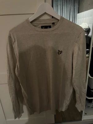 Beige stickad tröja Lyle & Scott M - Beige stickad tröja från Lyle & Scott i storlek M. Klassisk rund hals och långärmad modell med gul broderad logga på bröstet. Tröjan har ribbade muddar vid ärmslut och nederkant, perfekt för en clean och chill look.