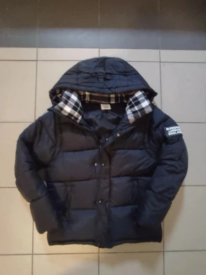 Svart Burberry pufferjacka  - Säljer en svart pufferjacka från Burberry med huva och klassiskt rutigt foder i svart, vitt och beige. Jackan har dragkedja och knappar framtill samt Burberry-logga på ärmen. Perfekt för kalla dagar och har en riktigt schysst streetstil. Ev byte