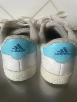 Vita Adidas sneakers med blå detaljer - Fräscha vita Adidas sneakers med klassiska blå ränder på sidorna och ljusblå hälparti med Adidas-logga. Skorna har snörning och platt sula, tillverkade i syntetmaterial och skinn. Perfekta för dig som gillar sportig och clean stil.