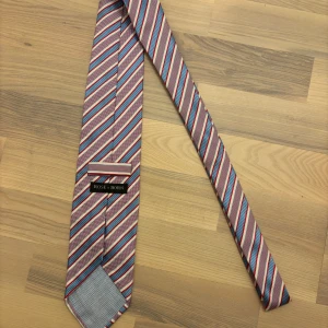 Randig sidenslips från Rose & Born - En elegant handgjord slips från Rose & Born, tillverkad i Italien av 100% silke. Slipsen har ett tidlöst mönster i rött, vitt och blått och är designad för att ge ett sofistikerat intryck vid både affärsmöten och formella tillställningar. Bredd: 9 cm, Längd: 150 cm. Kolla in min profil för mer liknande 😊 	