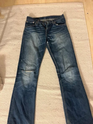 Levi's 501 blå jeans med slitning - Klassiska Levi's 501 jeans i blå denim med snyggt slitna detaljer och hål på ena knät. Rak passform och femficksmodell med knappgylf. Perfekta för dig som gillar en avslappnad och tidlös look.