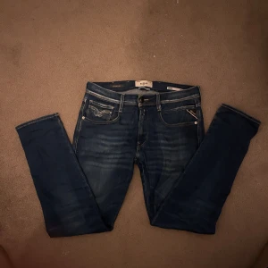 Replay Anbass blå jeans slim fit - Snygga Replay Anbass jeans i klassisk blå tvätt med diskreta slitningar och kontrastsömmar. Slim fit-modell med fem fickor och detaljerad bakficka. Perfekt passform och stilren look för dig som gillar moderna jeans.