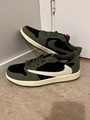 Travis Scott skor  - Nike Air Jordan 1 Low Travis Scott i olivgrönt och svart med vit omvänd swoosh. Skorna har röda broderade detaljer på hälen och klassisk snörning. Ovandelen är i skinn och mocka, med en platt sula och rund tå. Perfekt för dig som vill sticka ut med en ikonisk sneaker.