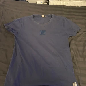 Blå t-shirt från SRNHLT, storlek 170 - Blå t-shirt från SRNHLT med rund hals och korta ärmar. Tryckt logga på bröstet och en liten patch längst ner. Skön passform och tillverkad i mjuk bomull, perfekt för dig som gillar enkel och clean stil.