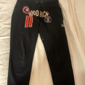 Svarta Hoodrich mjukisbyxor med tryck - Svarta mjukisbyxor från Hoodrich i storlek S med coola tryck och broderade detaljer framtill. Byxorna har snörning i midjan och flera patchar med logga och text. Perfekt för en streetwear-look och riktigt sköna att ha på sig.