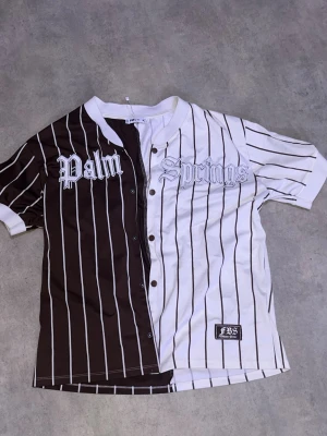 Randig baseballinspirerad topp Palm Springs - Säljer en unik baseballinspirerad topp från FBS Fashion Show med texten 'Palm Springs' broderad framtill. Halva toppen är brun och andra halvan vit, båda med tunna vertikala ränder. Kortärmad med knappar hela vägen och rundad hals.