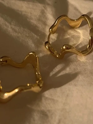 Guldiga vågiga örhängen från Pilgrim - Säljer ett par snygga örhängen från Pilgrim i guldtonad metall. De har en unik vågig och organisk form som ger en cool och modern vibe. Perfekta för att lyfta din stil med något annorlunda och trendigt. Kommer i originalförpackning.