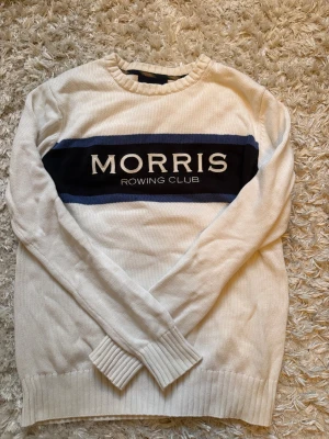 Vit stickad tröja Morris Rowing Club - Vit stickad tröja från Morris Rowing Club med mörkblått band och broderad logga över bröstet. Tröjan har rund hals, ribbade muddar och lång ärm. Perfekt för dig som gillar klassisk och sportig stil.