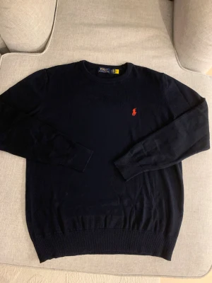 Mörkblå stickad tröja Polo Ralph Lauren - Snygg mörkblå stickad tröja från Polo Ralph Lauren i storlek XL men passar L. Klassisk rund hals och långärmad modell med ribbade muddar. På bröstet finns den ikoniska röda Polo-loggan broderad. Perfekt för dig som gillar stilrena och tidlösa plagg. 