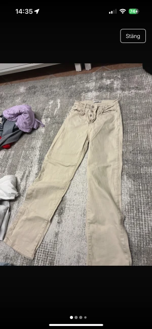 Beiga raka jeans från NA-KD, stl 34 - Snygga beiga jeans från NA-KD i rak modell med klassisk femficksdesign och knäppning med knappar framtill. Byxorna är i ett mjukt bomullstyg och har en stilren look med bakfickor och läderpatch med logga bak i midjan. Knappt knappt använda dock ser de skrynkliga ut på bild då de är nytvättade. 