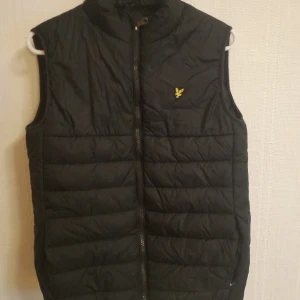 Svart dunväst från Lyle & Scott Junior - Svart dunväst från Lyle & Scott Junior med gul logga på bröstet. Västen har quiltad design, hög krage och dragkedja framtill. Perfekt för lager-på-lager och har en stilren look som funkar till många outfits.