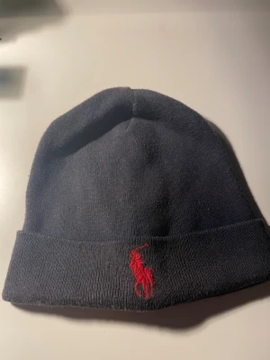  Ralph Lauren mössa  - Snygg mörkblå mössa från Polo Ralph Lauren med klassisk röd broderad logga framtill. Mössan är stickad och har en uppvikt kant som ger extra stilpoäng. Perfekt för att hålla värmen och samtidigt se fräsch ut under kyliga dagar.