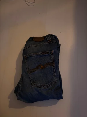 Blå jeans från Nudie  - Säljer ett par klassiska blå jeans från Nudie med snyggt brunt broderi på bakfickorna och läderpatch i midjan. Jeansen har rak passform och är tillverkade i slitstark bomull. Perfekt för dig som gillar en tidlös look och vill ha jeans med coola detaljer. Jag har ett hål precis i röven och ett på knäskålen därför priset men det är lätt att fixa! W29 L32