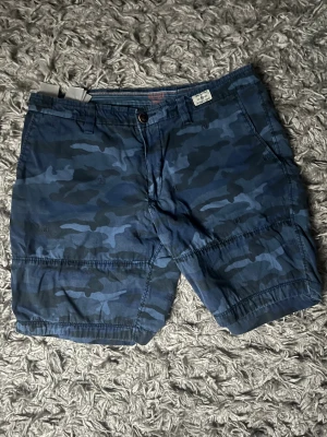 Blå camo shorts från Tommy Hilfigher - Säljer ett par gamla blå camouflage-mönstrade shorts från Tommy hilfigher. De är i storlek 31 och är i regular fit. Dem är i ett bra skick.