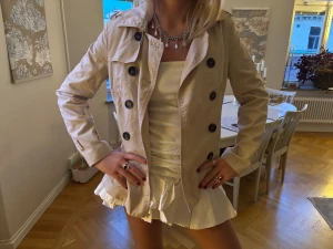 Beige trenchcoat med stora knappar - Snygg beige trenchcoat med klassisk krage och stora svarta knappar framtill. 