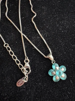 Blomformat halsband i äkta silver - Snyggt halsband i äkta 925 silver med en blomformad hängare dekorerad med turkosa stenar. Kedjan är tunn och har en justerbar längd med karbinlås. Perfekt för dig som gillar unika detaljer och färg. Blomman har fem kronblad och gnistrar fint i ljuset.